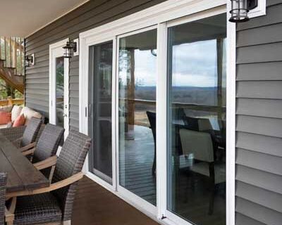 Outdoor deck with sliding glass doors, wicker chairs, and a wooden table beside a house with gray siding.