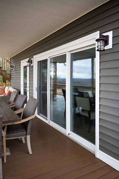 Outdoor deck with sliding glass doors, wicker chairs, and a wooden table beside a house with gray siding.