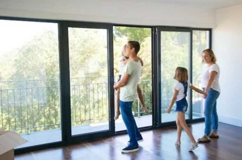 Group of people standing inside a room with large floor‑to‑ceiling sliding glass doors overlooking greenery.