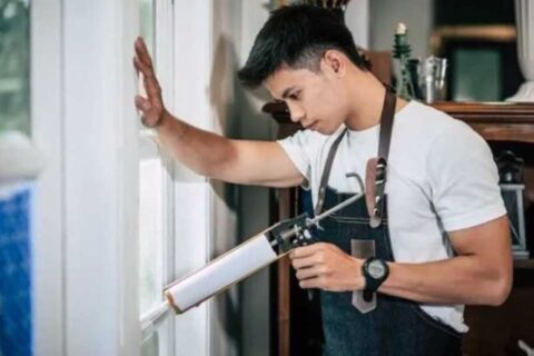 Person applying sealant to a window frame while wearing an apron and holding a caulking gun.