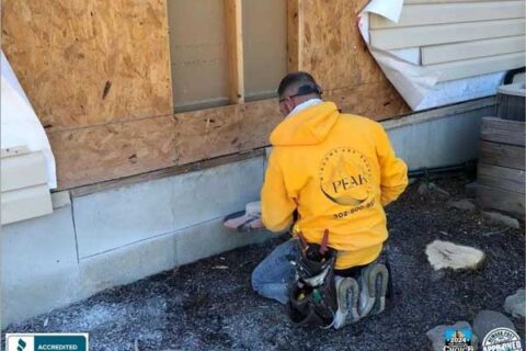 Worker in a yellow Peak Windows and Doors hoodie repairing the exterior wall of a house.