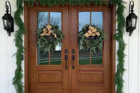 Double wooden front doors decorated with evergreen garlands, wreaths, and flanked by black lantern-style wall lights.