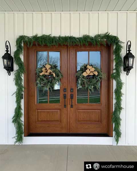 Double wooden front doors decorated with evergreen garlands, wreaths, and flanked by black lantern-style wall lights.