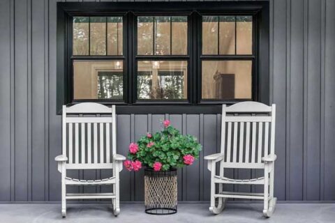 Front porch with two white rocking chairs and a potted flower arrangement in front of a black‑trimmed window.