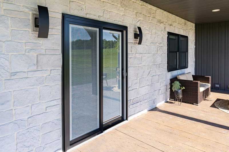 Sliding glass patio door with built-in blinds on a stone exterior wall beside a small outdoor seating area.