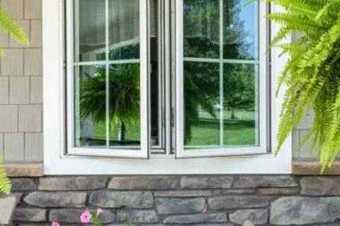 Tilted double-hung window with white framing above a stone exterior wall, surrounded by green plants.