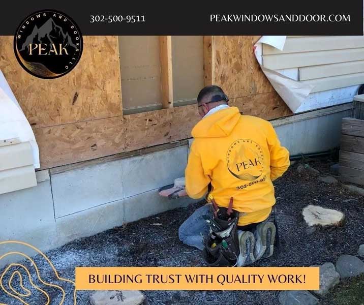 Worker in a yellow Peak Windows and Doors hoodie applying material to the exterior foundation of a house under renovation.