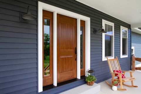 Front porch with a wooden door, glass sidelights, navy siding, and a wooden rocking chair with a red pillow.