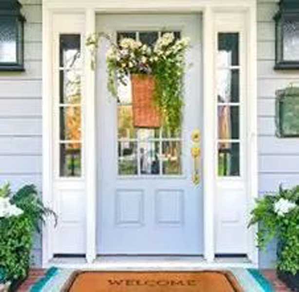 Front door with sidelights, floral basket décor, welcome mat, and potted plants on each side.